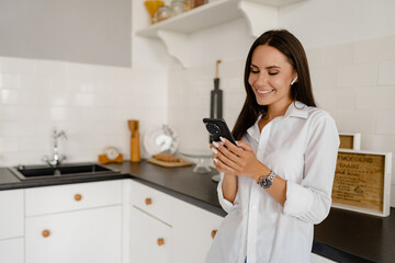 woman in kitchen at home relaxed using smartphone in white t-shirt and jeans listening to earpods