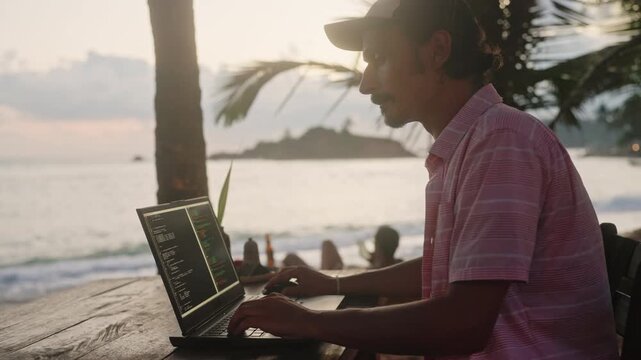 Software dev works on laptop at beachside cafe. Man writes code during ocean sunset, palm trees sway. Freelance programmer creates software near sea, island view in background. Slow motion.