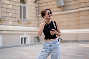 Fototapeta premium stylish woman with short haircut walking in street with coffee wearing black top, jeans and sunglasses accessories