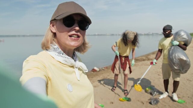 Medium selfie shot of Caucasian woman in baseball cap filming herself on smartphone speaking about beach litter pick community event, demonstrating diverse team collecting rubbish, waving at camera