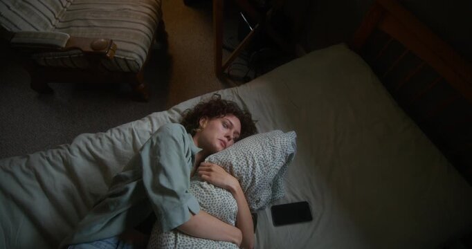 A woman is crying and hugging a pillow lying in bed at home. The mascara is smeared under the eyes. The mobile phone is on her bed