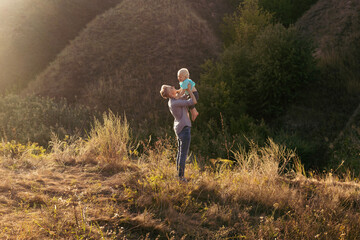 Mother and her little son enjoy they time in nature in summer with sun rays on sunset near green hills