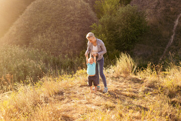 Happy young mother lifts up her son while playing outdoors in nature in summer with sun rays on sunset near green hills