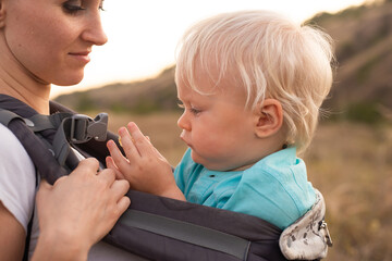 Mother carries her little baby boy in a sling in summer nature with sun rays on sunset