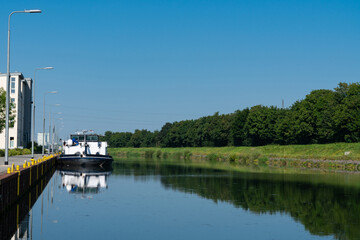 Obraz premium A barge rests quietly by the canal, reflecting the clear blue sky and surrounding greenery in the calm water.
