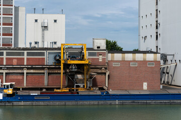 A yellow loading crane is busy transporting goods at an industrial dock beside a tranquil waterway surrounded by urban buildings.