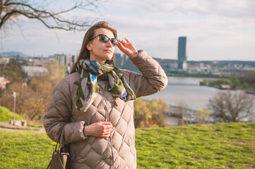 Young beautiful woman in sunglasses, coat, and jeans stands on a hill with a view of Belgrade, Serbia