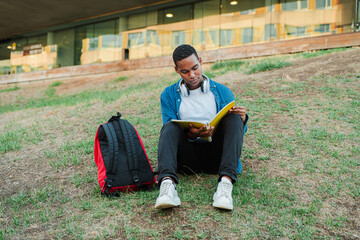 African american handsome guy reading a note book to prepare a exam. Teenage student sitting on the university campus lawn and studying a class lesson. Young man doing the homeworks for the academy