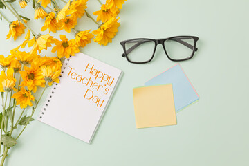 Fat lay composition with notebook with text "Happy Teacher's Day", yellow daisy flowers, and black stylish glasses, sticker note on a pale light green background. Top view with copy space