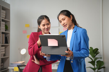Two businesswomen are standing together in their office, reviewing work on a digital tablet