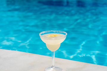 A margarita cocktail with lemon sits on the edge of a swimming pool