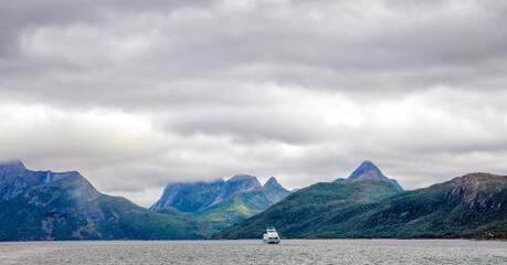 Small ferry in an ocean with remote Lofoten islands dramatic peaks on background. Cloudy sky....