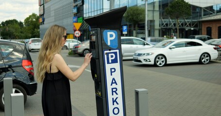 Young woman in sunglasses and summer dress stand at parking meter and enter code. Beautiful female...