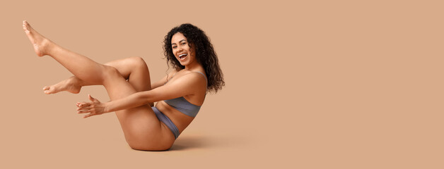 Beautiful young happy African-American woman in stylish underwear sitting on beige background