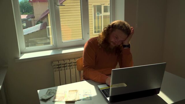 bearded man working in an office, he gets a headache and grabs his head but continues to work on his laptop