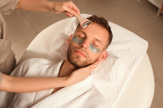 Handsome young man in under-eye patches receiving facial massage with roller in beauty salon