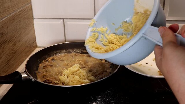 A woman transfers pasta risoni from a colander to a frying pan with mushroom sauce. The process of preparing mushroom dishes.