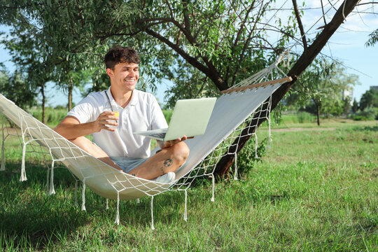 Young man with lemonade and laptop resting in hammock outdoors