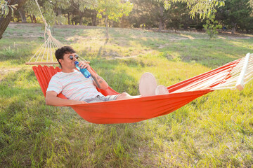 Young man in sunglasses with bottle of water resting in hammock outdoors