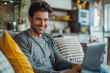 Fototapeta premium The beautiful young man is smiling and looking at his laptop while sitting on the couch.
