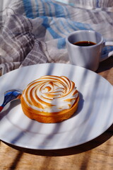 Appetizing beautiful lemon tartlet with meringue on a white plate, on a stylish wooden background with a blue-gray coton, next to a cup of coffee The best typical French baking
