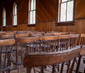 Simple old wooden chairs in a plain United Church with arched windows in the background.
