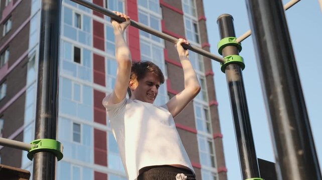 Weak funny teenager strains in an attempt to pull himself up on the horizontal bar