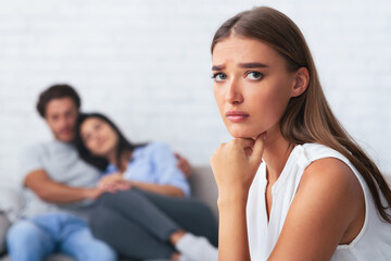 Jealousy. Sad Girl Looking At Camera While Her Ex-Boyfriend Hugging New Girlfriend Sitting On Couch...