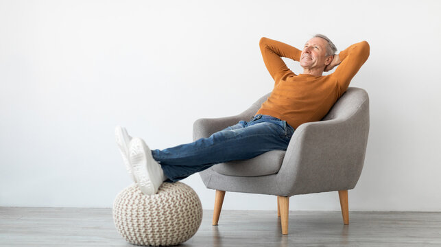 Smiling calm mature man relaxing sitting on armchair, resting feet on knitted pouf, free copy space. Happy adult spending weekend, leaning back holding hands behind head, isolated on white studio wall