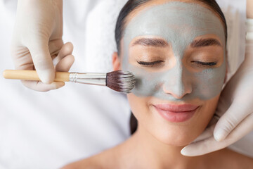 Aesthetic procedure. Beautician applying grey face mask on young female face, woman enjoying anti-aging skincare treatment