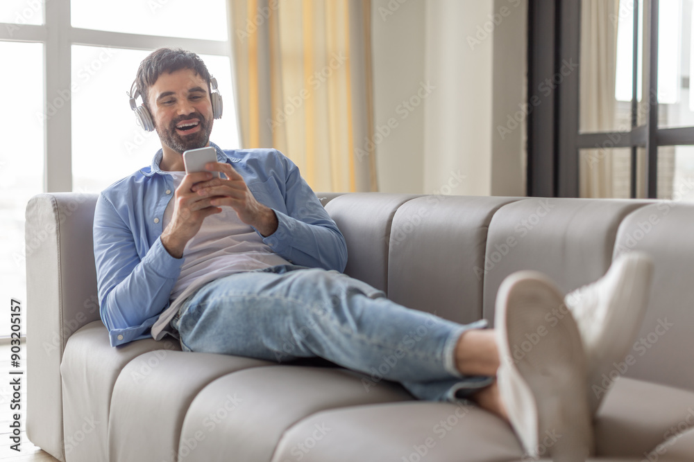 Poster a man is lying on a grey couch with his legs extended and wearing headphones. he is smiling and look
