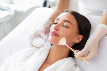 Cosmetologist cleaning face of a beautiful European woman with special gel before applying mask,...