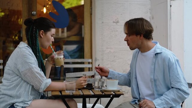 A young Caucasian couple on a date in a summer cafe. A guy and a girl are talking, laughing, drinking coffee and eating delicious desserts on the terrace of the coffee shop