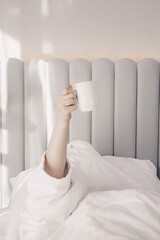 A sleepy young woman with cup of coffee lying in bed with white cotton bedclothes. Concept of early morning wake up. Pastel colors. Minimalism. Sunday loneliness relaxing, hygge mood.
