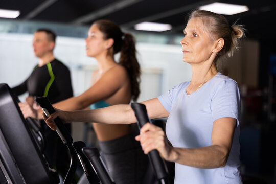 Portrait of motivated sporty senior woman doing cardio training, exercising on elliptical trainer in gym