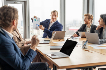 A group of business professionals are gathered around a table in an office. One man is presenting data from a document to the group while the other colleagues listen attentively, engage in discussion.