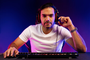 Smart gaming streamer looking at camera with trendy pose, passing the level of online game user on pc, using headset and mic for communicate with others at digital neon light studio room. Surmise.