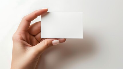 Obraz premium A close-up of a hand holding a blank white business card against a soft white background, ideal for customization and branding needs. 