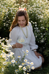 Woman with a glass in her hands is sitting in a chamomile field. Girl in daisies