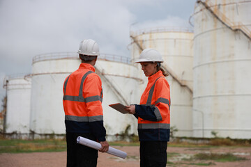 Fototapeta premium The senior manager female engineer and her male colleague are confidently working together at an oil storage tank field with a blueprint, utilizing a tablet to effectively track the progress of the pr