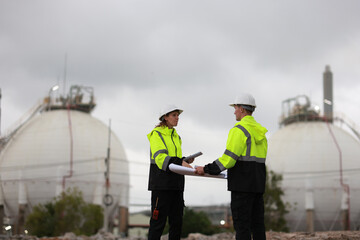 Male & Female engineers stand Discussing the building plan at the site, Engineers working outdoors and use walkie-talkies against the Round gas storage tank in the background