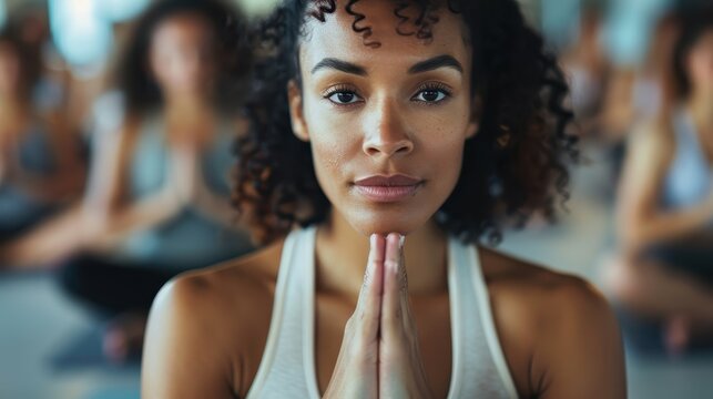 A woman in a yoga class has her hands in prayer pose, with an expression of calm and focus, surrounded by other participants blurred in the background.