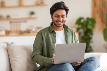 Arab man is sitting on a white couch in a modern home. He is smiling and looking at his laptop. He...