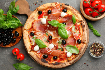 Tasty pizza with cured ham, olives, mozzarella cheese, tomatoes and arugula on gray table, flat lay