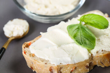 Piece of bread with cream cheese and basil leaves on gray table, closeup