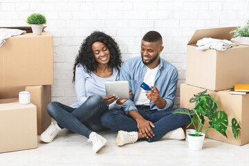 Young black couple buying furniture online, using digital tablet and credit card, sitting on floor in new apartment