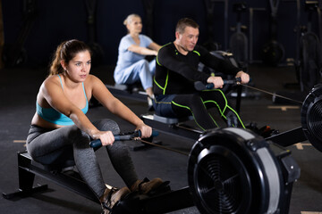 Portrait of concentrated young girl doing intense full body workout in gym, exercising on rowing machine