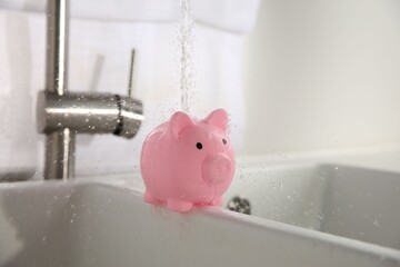Water saving concept. Stream of water flowing from tap onto piggy bank in kitchen