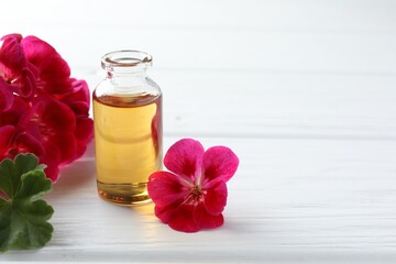 Bottle of geranium essential oil and beautiful flowers on white wooden table. Space for text