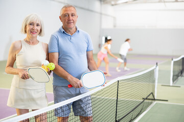 Experienced team of pickleball players, friendly smiling aged woman and man, standing on indoor court with paddles and balls in hand ready to training